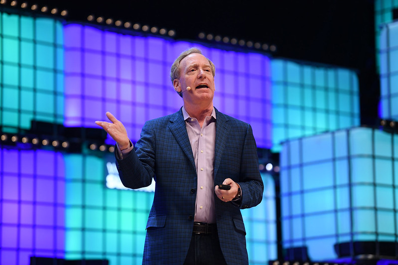 A photograph of a person, Microsoft president Brad Smith, speaking on Centre Stage at a Web Summit event. They are gesturing with their hands and wearing an on-ear microphone.