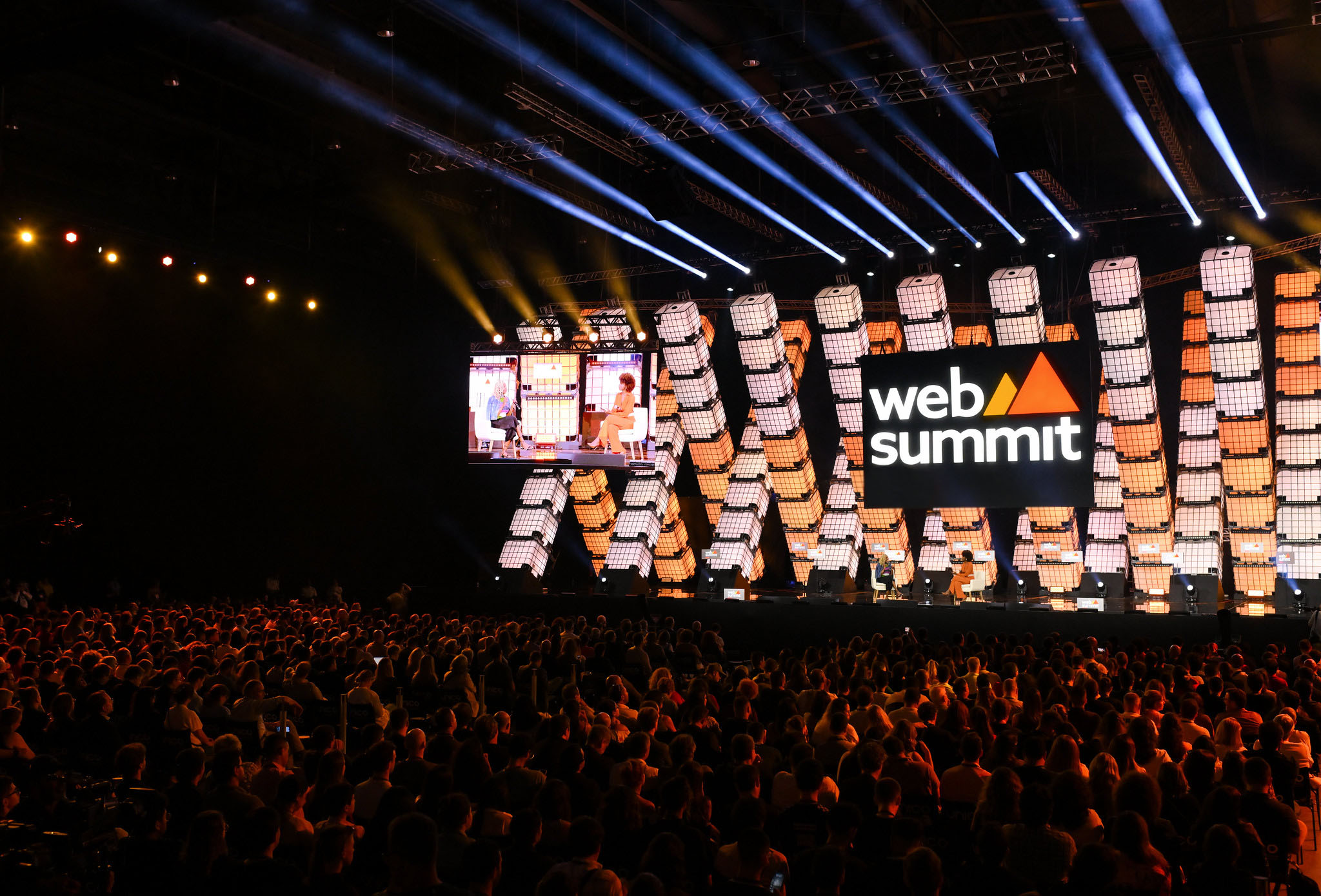 A zoomed-out photograph of Center Stage during the Opening Night at Web Summit Rio. There is a large audience of people sitting in front of a lit-up stage, with the Web Summit logo front and centre.