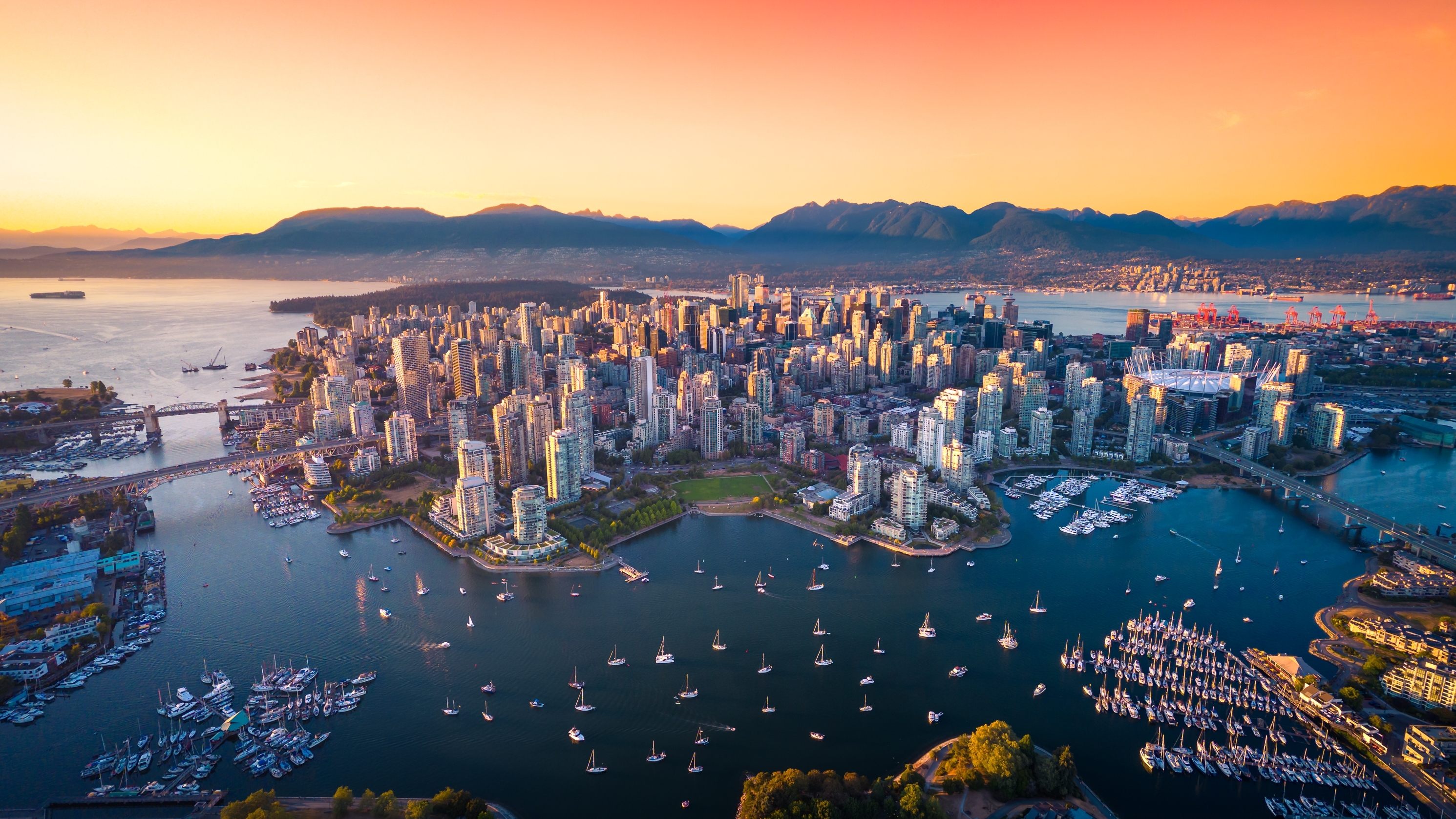 A birds-eye image of Vancouver. The city sits in the middle of the bay, with mountains visible in the background. The photo appears to be taken at sunset.