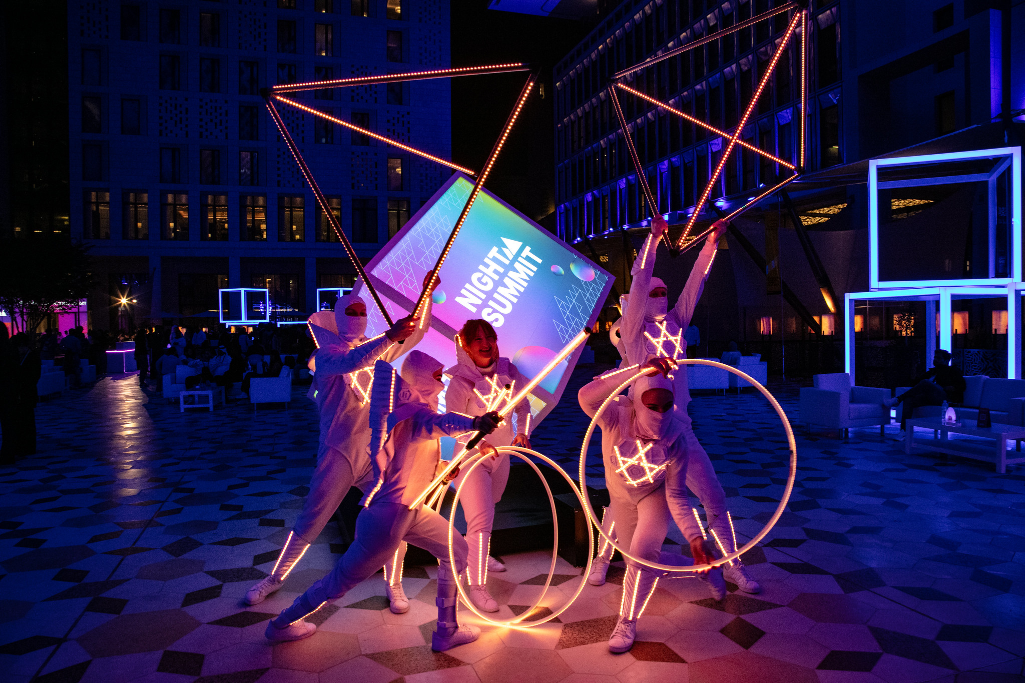 A photograph of a live art installation at Night Summit in Qatar, at Barahat Msheireb. There are five people holding up 3D shapes, dressed in all-white suits. There are light installations visible in the background.